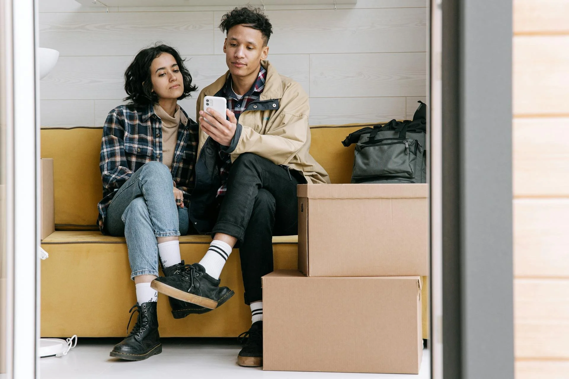 Couple sitting on a yellow couch and reviewing property options on a smartphone beside moving boxes